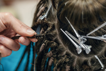 Process of braiding dreadlocks, close-up. Human dark brown hair, hairdressing clips and hairdressers...