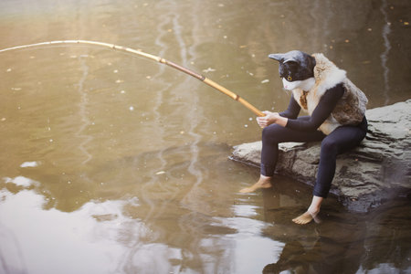 A person in quadrober costume in a cats mask and fur vest sits on a rock on the riverbank with a bamboo fishing rod in his hands.の写真素材