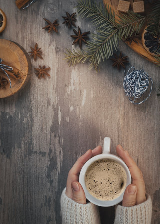 Christmas flat lay. Female hands in a cozy sweater with coffee cup and spruce twigs, star anise, brown sugar, a ball of blue twine on a wooden background, vertical image.の写真素材