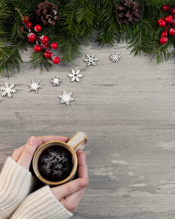 Christmas flat lay. Female hands in a cozy white sweaters sleeves with coffee cup and frosty patterns in it. Tradition border with green spruce, cones, holly berries.の写真素材