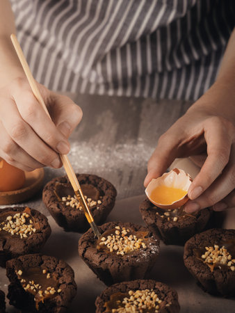 Bakers hands grease the muffins with a brush with egg yolk on a table, close-up, vertical image.