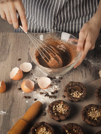 Female hands beat with a whisk dough into a bowl next to muffins, eggshell and rolling pin on on a table, faceless baker in striped apron, vertical image.の写真素材