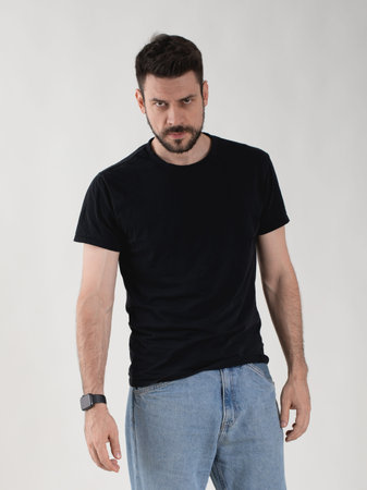 Portrait of a 35 years old bearded man in a basic black t-shirt and blue jeans. He looks directly at...