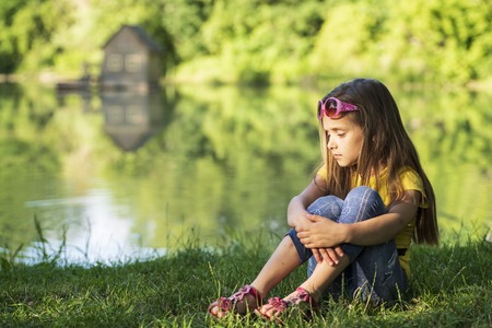 The girl is sad on the river bank, on a sunny summer day.The little girl sitting on the river bank, having covered her face with her hands,の写真素材