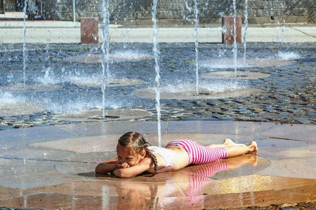 Girl on a sunny summer day are poured water from a fountain.Girl happily in shallow clean water on of city fountain on warm bright summer day.の写真素材