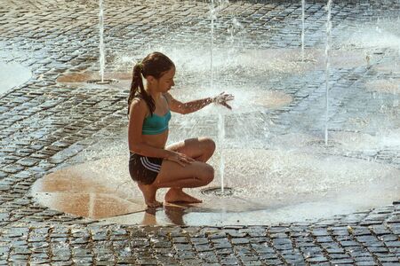 Girl on a sunny warm day playing outside in a water fountain. Girl happily in shallow clean water on of city fountain on warm bright summer day.の写真素材