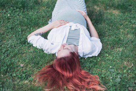 Pregnant white girl lies on green grass in a light striped dress on a summer day. Girl expecting a baby. Pregnant girl in the park on a summer day.Closeup pregnant young woman outdoors in warm summer dayの写真素材