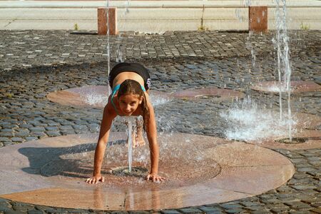 Girl on a sunny summer day are poured water from a fountain.Girl happily in shallow clean water on of city fountain on warm bright summer day.の写真素材