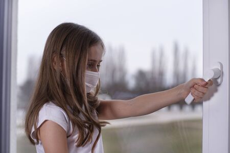 A little girl sits at the window in a protective mask, wants to go outside, but it s impossible, there is a coronavirus, the girl is sad. Girl in a protective mask by the window.の写真素材
