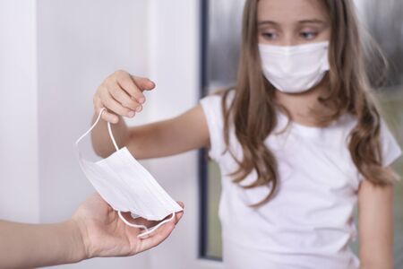 Personal protective equipment required.Coronavirus.A little girl sits by the window in a protective mask, offers to put a protective mask on people and protects herself from the virus.の写真素材