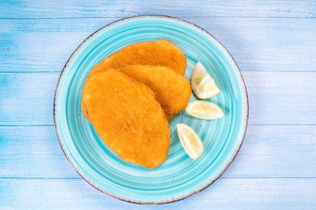 Breaded chicken schnitzel on a blue plate with slices of lemon on a blue wooden background.Top view,...