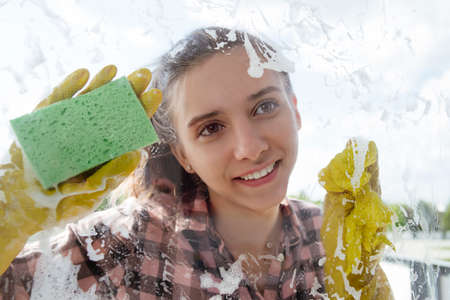 Cleaning house conceptA young girl with a smile in yellow rubber gloves washes a window from the outside.の写真素材