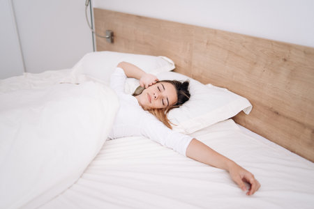She keeps eyes closed and looks satisfied.Portrait pretty young girl on bed in modern apartment in the morning.の写真素材