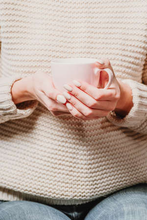 A girl in a beige sweater holds a cup of tea in her hands.の写真素材