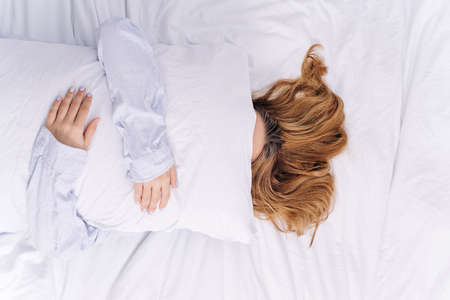 Young girl lying in bed covering her face with white pillow. Concept of awakening, lazy day.の写真素材