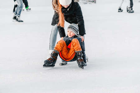 Soft,Selective focus.Mom with baby boy 6 years old, learn train, ride winter city rink, ice skating. The child gets up, fell on skates, kneels, play fun rest on weekend first steps child skatesの写真素材