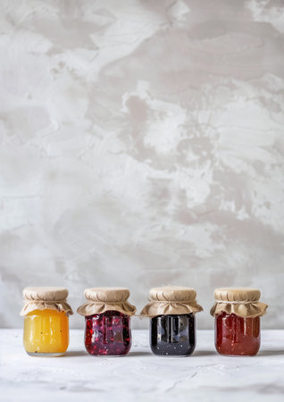 Four colorful glass jars with fabric lids and jam of different flavor and color on marble surface and white background. High quality photoの素材