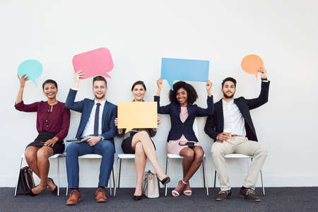 Studio portrait of a group of young businesspeople holding speech bubbles while sitting in a row against a white backgroundの写真素材