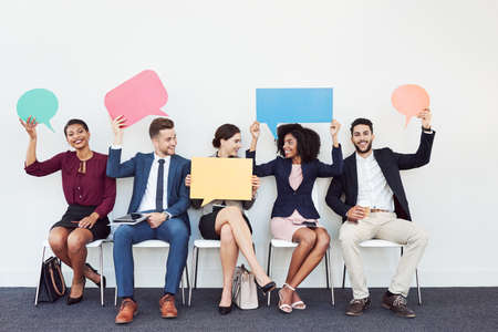 Studio shot of a group of young businesspeople holding speech bubbles while sitting in a row against a white backgroundの写真素材
