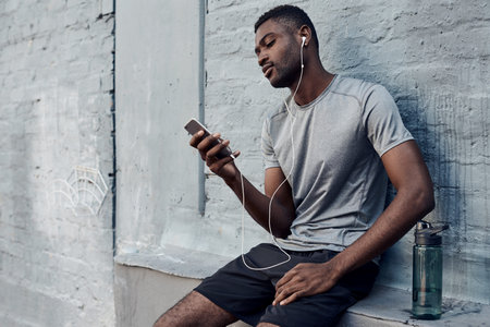 Shot of a sporty young man wearing earphones while using a cellphone against a grey wall