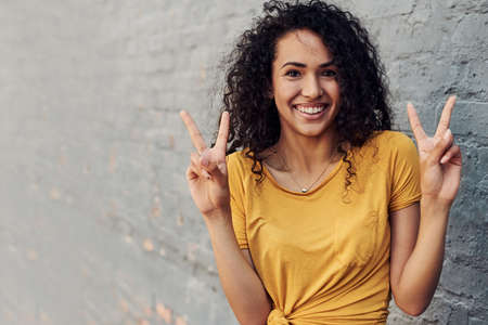 Cropped portrait of an attractive young woman standing alone outside and making peace sign hand gestures against a gray backgroundの写真素材