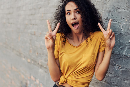 Cropped portrait of an attractive young woman standing alone outside and making peace sign hand gestures against a gray backgroundの写真素材