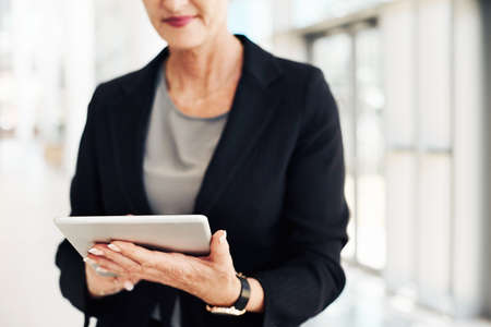 Cropped shot of an unrecognizable mature businesswoman using a digital tablet while standing in a modern officeの写真素材