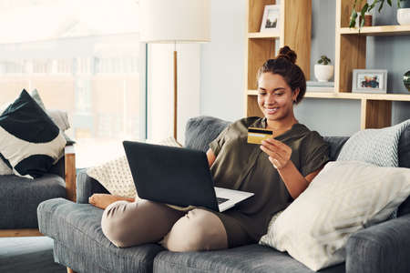 Shot of a young woman using a laptop and credit card on the sofa at homeの写真素材