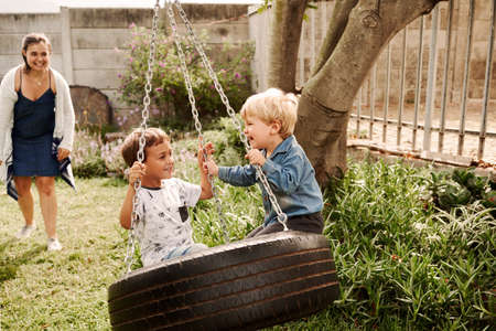 Shot of two adorable little boys playing on a swing together outdoors with their mother watching in the backgroundの写真素材