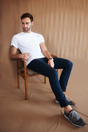 Shot of a handsome young man sitting on a chair against a brown backgroundの写真素材