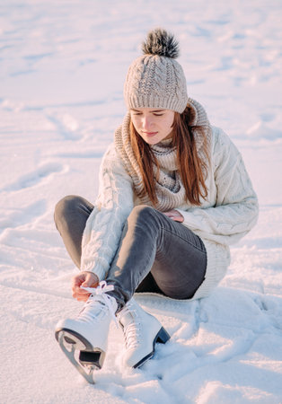 Young woman ice skating on a frozen lake in a snowy winter park during sunset. A girl sits on the ice tying her skates before skating at the skating rink. Active recreation in winter.の写真素材
