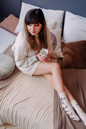 Young woman holding a cup of tea on a winter morning. Dreaming girl sitting on the bed with a cup of hot coffee, enjoying at home. Year holidays concept - woman with a cup of cocoa in bed at home.の写真素材