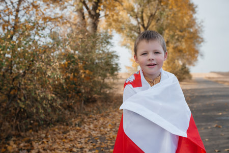 A handsome little boy is wrapped in the flag of Poland. Portrait of a child with a national flag in nature. Concept of national holidays and patriotic events. Patriotism. Independence Day.の写真素材