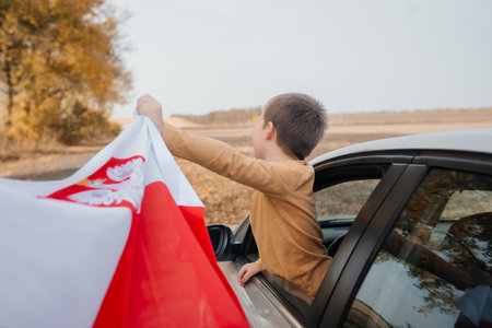 A little boy in a brown sweater in nature holds the flag of Poland in his hand. A child looks out of the car waving a flag. Rear view. Concept of national holidays and patriotic events. Patriotism.の写真素材