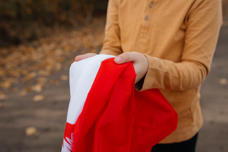 A little boy stands on the road and holds the flag of Poland in his hands. Close-up of child's hands in brown sweater with flag. Concept of national holidays and patriotic events. Independence Day.の写真素材