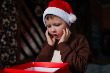 A child in a red Christmas hat opens a gift box received for Christmas and makes an excited gesture. Portrait of amazed little boy looking inside red box unwrapping gift for New Year.の写真素材