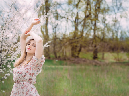 A woman in a white dress with a colorful pattern enjoys the aroma of flowers. A girl is standing near a blooming spring tree in the garden with her arms raised. Background with space for text.の写真素材