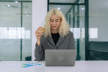 Businesswoman sitting and looking at data on laptop. Woman drinking in front of computer in office.の写真素材