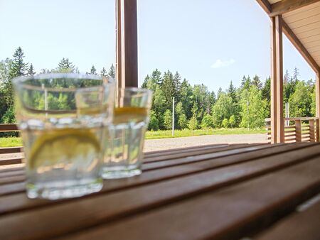 Wooden country furniture on the terrace of a country house. Two refreshing drinks out of focus with a refreshing lemonade with lemon slices in the foreground. Lush green foliageの写真素材