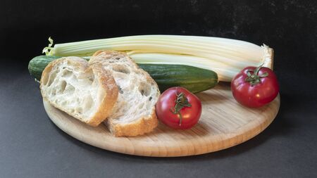 Two slices of white beautiful airy white bread, fresh vegetables - cucumbers, tomatoes and celery, all on a round wooden cutting board on a black background.の写真素材