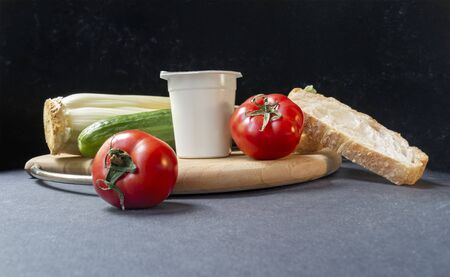 Slices of white beautiful airy white bread, yogurt, fresh vegetables - cucumbers, tomatoes and celery, all on a round wooden cutting board on a black background close-up, side view.の写真素材