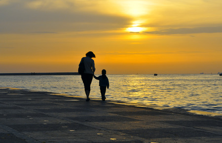Silhouette of a woman with a small child, they hold hands on a background of sea sunset on the dockの写真素材