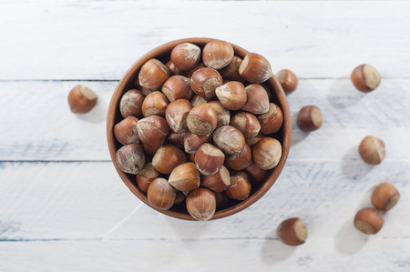 Full bowl of hazelnuts in a brown bowl, top view, white wooden backgroundの写真素材