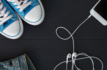 Smartphone with headphones and blue sneakers on a black wooden surface, top view