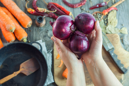 female hands holding three red onions on the background of the table with vegetables and spices, near black cast iron pan with a wooden spatula, top viewの写真素材