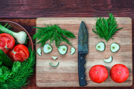 Fresh tomatoes and cucumber in a bowl and on a kitchen board, funny faces males and women from pieces of cucumber and a bunch of dill, top viewの写真素材