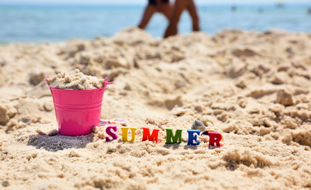 Inscription summer on the sand against the blue sea, next to a full pink bucket of sandの写真素材