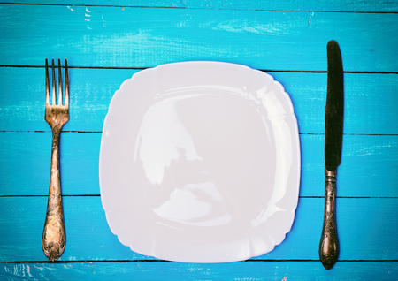 Empty square white plate with iron fork and knife on a blue wooden background, top view