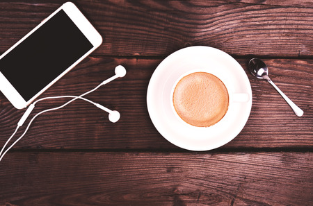 white cup with coffee and foam, side view is a white smartphone with headphones on a brown wooden background, top viewの写真素材