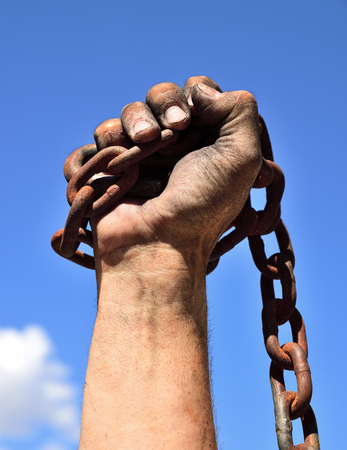 man's hand wrapped in an iron rusty chain lifted up against a blue skyの写真素材
